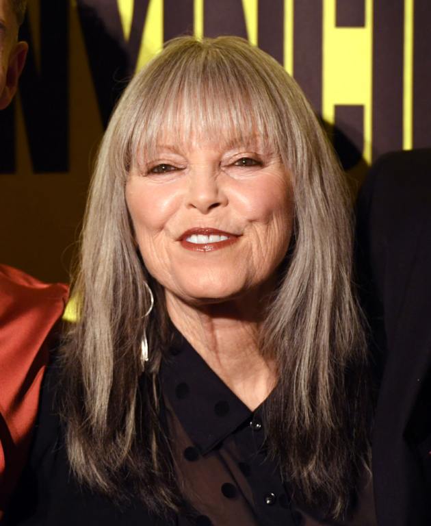 Pat Benatar smiling at the opening night for the world premiere production of "Invincible: The Musical" wearing a black mesh polka dot button up and long grey hair with bangs and a red lip.