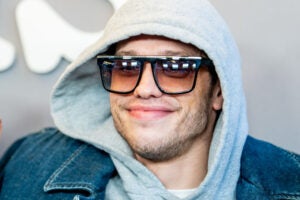 Pete Davidson attends the Peacock's "Bupkis" World premiere smiling facing left wearing sunglasses, grey hoodie, and denim jacket.