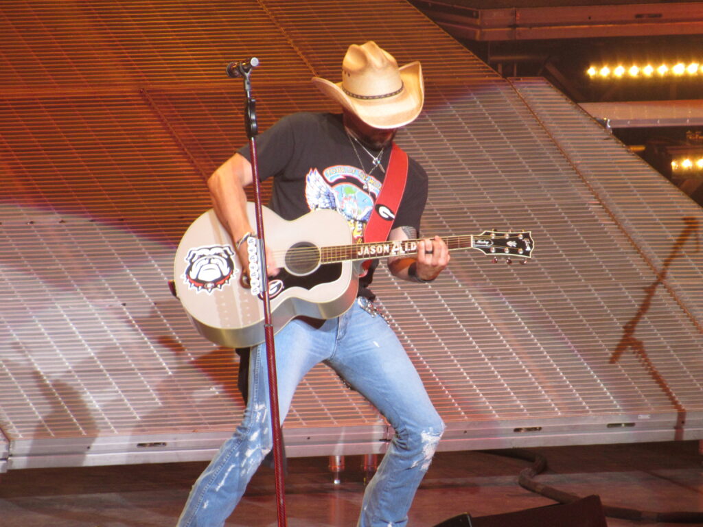 Jason Aldean leaning back while playing an acoustic guitar on stage in 2018