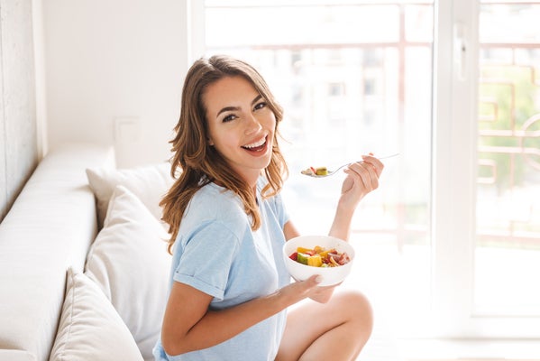 young brunette eating a bowl of cereal and laughing on the couch