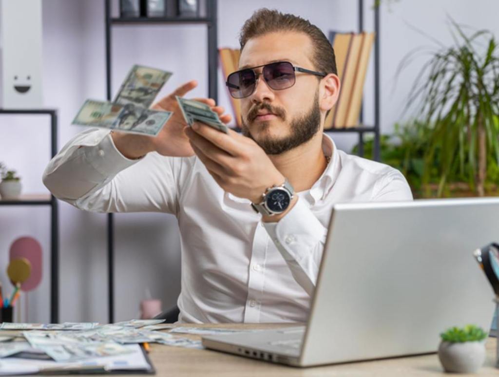 Cheerful rich business man working on office laptop wasting throwing money to camera profit win (Redditors Share Their Weirdest Money-Making Stories)