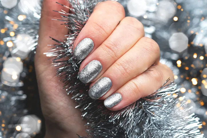 Female hand with beautiful holiday manicure - silver glittered nails on Christmas tinsel background with blurred lights