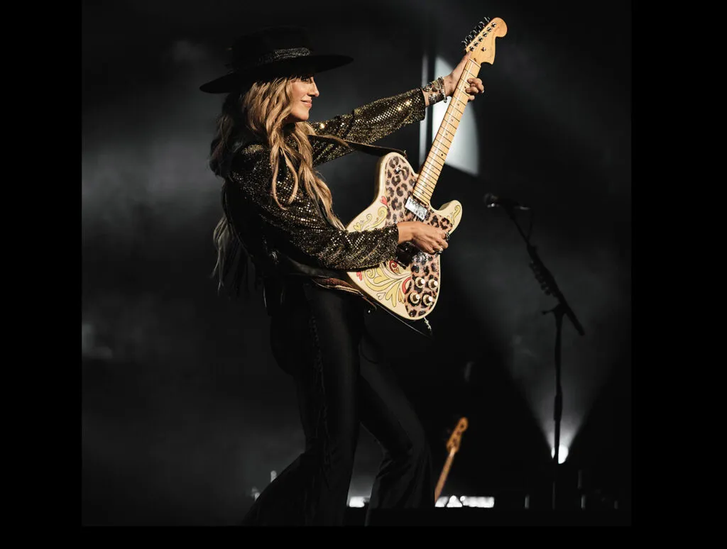 Black background with Lainey Wilson in the middle in dark clothing, playing guitar