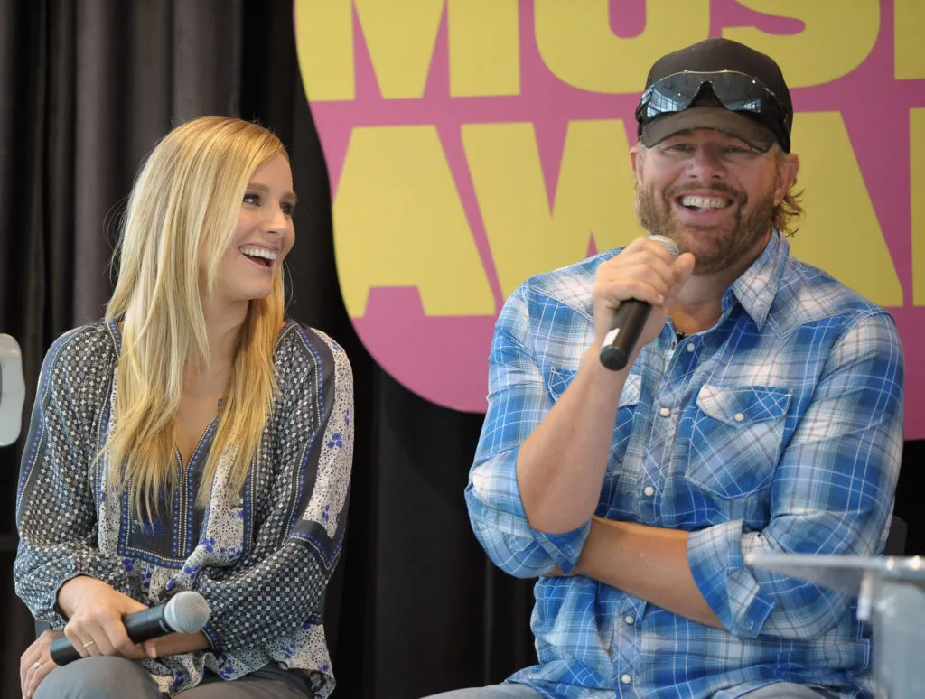 CMT Will Pay Tribute To Toby Keith - Toby in 2012 at a press conference with CMT co-host Kristen Bell before the CMT Awards. Toby is in a plaid shirt, and Kristen is wearing a gray outfit.
